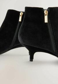 Stivaletti neri in suede con un design elegante, caratterizzati da dettagli dorati, zip laterali e un tacco a kitten basso.