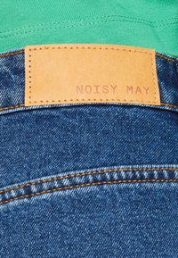Denimkangas sinisellä pinnalla, jossa on oranssit ompeleet ja vaaleanruskea nahkapaikka, johon on merkitty "NOISY MAY" vyötäröllä. Vihreä kangas näkyy ylhäällä.