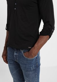 Zwarte langarm henley-shirt met drie knopen bij de halslijn en opgerolde mouwen, gecombineerd met versleten blauwe spijkerbroek.