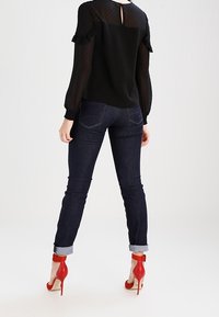 Sort bluse med polka prikker i gennemsigtige ærmer, parret med mørke denim jeans rullet op ved anklerne, og røde højhælede sko med ankelstropper.
