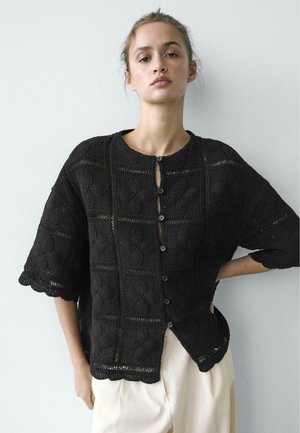 Jonge vrouw draagt een loszittend zwart gehaakt blouse met knopen en ellebooglange mouwen, en witte broek, staand tegen een eenvoudige lichte muur.