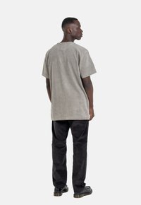 Kurzärmliges, hellgraues, samtiges T-Shirt mit lockerer Passform, kombiniert mit schwarzen, strukturierten Hosen und dunklen Schuhen, von hinten gesehen.