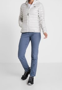 Jasnoszara kurtka puchowa z pikowaną fakturą i kapturem, w zestawie z niebieskimi spodniami i czarnymi butami do wędrówek, z sznurówkami i gumową podeszwą.