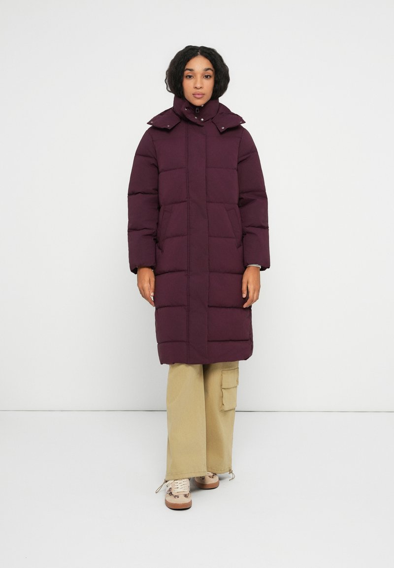 Kvinde iført en lang lilla polstret frakke med høj krave, beige cargo-bukser og beige sneakers, stående foran en helt hvid baggrund.