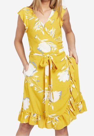 Kvinde iført en gul slå-om kjole med hvidt blomsterprint, korte ærmer, flæsekant og bindebælte i taljen, hænderne hviler i lommerne.