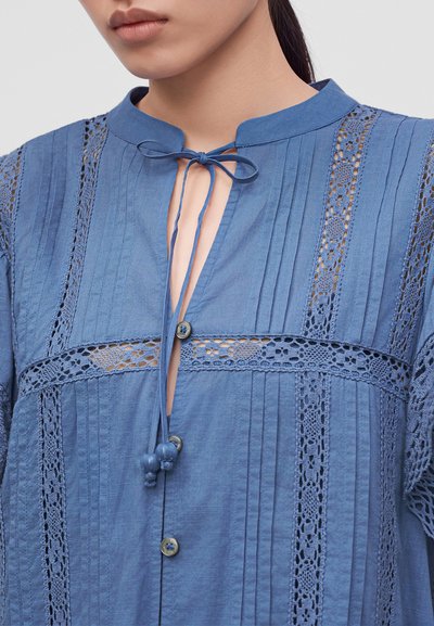 Femme portant un chemisier bleu avec des panneaux en dentelle, des plis verticaux, des boutons et un cordon noué au cou avec des perles décoratives.