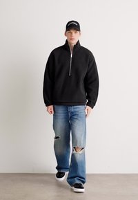 Un tânăr îmbrăcat cu un fleece negru cu fermoar pe jumătate, blugi rupți albaștri, adidași negri și o șapcă neagră, stând în fața unui perete alb simplu.