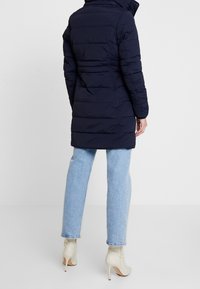 Cappotto imbottito blu navy con colletto alto, maniche lunghe e motivo a righe orizzontali, abbinato a jeans blu chiaro e stivaletti beige.