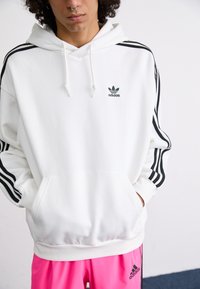 Sweatshirt branco com bolso canguru, riscas pretas nas mangas e pequeno logótipo preto da Adidas no peito. Acompanhado de calções rosas.