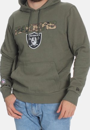 Homme portant un sweat à capuche Raiders vert olive avec texte camouflage, logo de l'équipe, patch NFL sur la manche, et jean bleu clair.