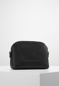 Malá černá texturovaná kožená taštička se zipovým uzavřením, umístěná na bílé ploše proti jednoduchému světlem pozadí.