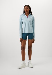 Light blue long-sleeve top with a zip collar, paired with teal shorts, white sneakers, and white socks, featuring a smooth texture and athletic design.