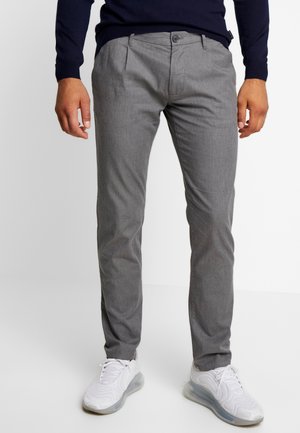 Homme portant un pantalon gris ajusté et des baskets blanches, debout devant un fond blanc uni.