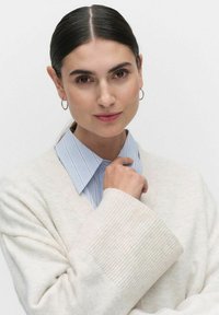Krämsfärgad cardigan med ribbade manschetter, lagd över en ljusblå och vit randig skjorta. Modellen har rakt svart hår och hoopsörhängen.