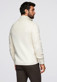 Mężczyzna z ciemnymi włosami i brodą, ubrany w biały sweter w warkocze i ciemne spodnie, lekko zwrócony w bok na jednolitym tle.