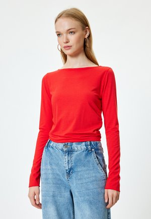 Rød langærmet bluse med bådudskæring, parret med lyseblå højtaljede jeans. Stoffet ser blødt og tætsiddende ud.