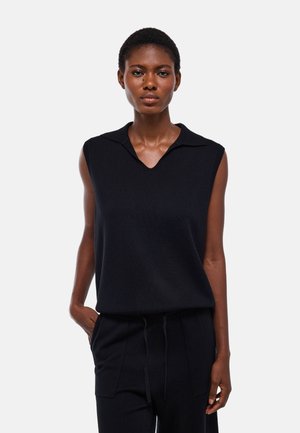 Femme portant un haut noir en maille sans manches avec col et un pantalon noir, debout devant un fond uni clair, une main dans la poche.