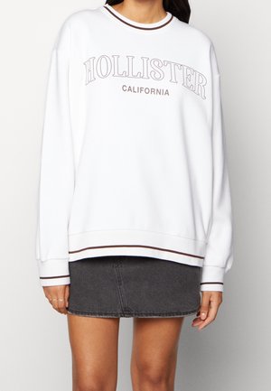 Sweatshirt blanc avec le texte "HOLLISTER CALIFORNIA" en brun, présentant un col rond, des manches longues, et des poignets et un ourlet rayés.