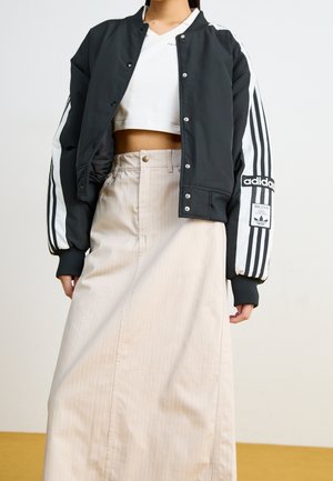 Osoba nosící černou bomber bundu Adidas s bílými pruhy, bílý krátký top a béžovou sukni s vysokým pasem.