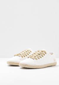 Bílé plátěné tenisky se zlatými třpytivými tkaničkami, pletenou juteovou podešví a nízkým profilem. Žádné viditelné logo nebo vzory.