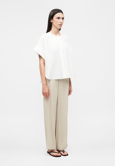 Γυναίκα που φοράει λευκό άνετο μπλουζάκι με κοντά μανίκια, μπεζ παντελόνια με φαρδιά πόδια και μαύρα σαγιονάρες, στέκεται μπροστά σε απλό φωτεινό φόντο.
