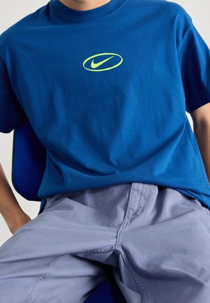 Personne portant un t-shirt Nike bleu avec un logo vert fluo et un pantalon violet clair, assise avec les mains reposant sur les cuisses.