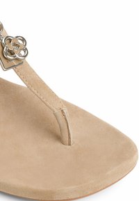 Sandalo infradito in suede beige con un accento metallico floreale sulla cinghia, dotato di un plantare liscio e arrotondato e di un design minimalista.