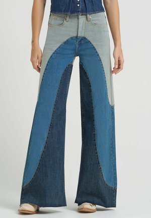 Žena oblečená v džínsach s vysokým pásom a širokými nohavicami, ktoré majú svetlomodré a tmavomodré zakrivené záplaty, a nosí béžové topánky.