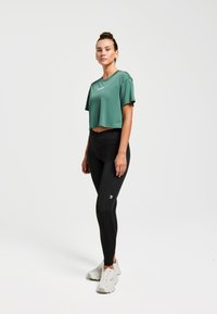 T-shirt athlétique vert court, associé à un legging noir taille haute. Baskets blanches avec des semelles texturées, présentant un petit logo sur le legging.