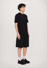 Melna kokvilnas T-krekls ar īsām piedurknēm un krūšu kabatu, saskaņots ar brīviem melniem šortiem. Modelis nēsā melnus slip-on apavus un baltas zeķes.