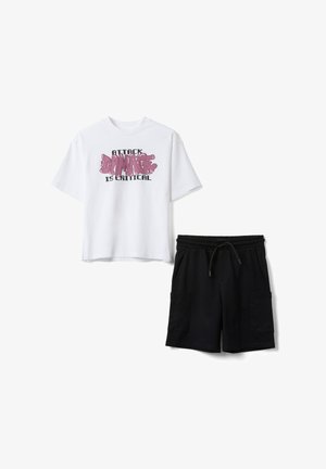 T-shirt blanc à manches courtes avec le texte « Attack Damage is Critical » associé à un short cargo noir à cordon sur fond blanc.