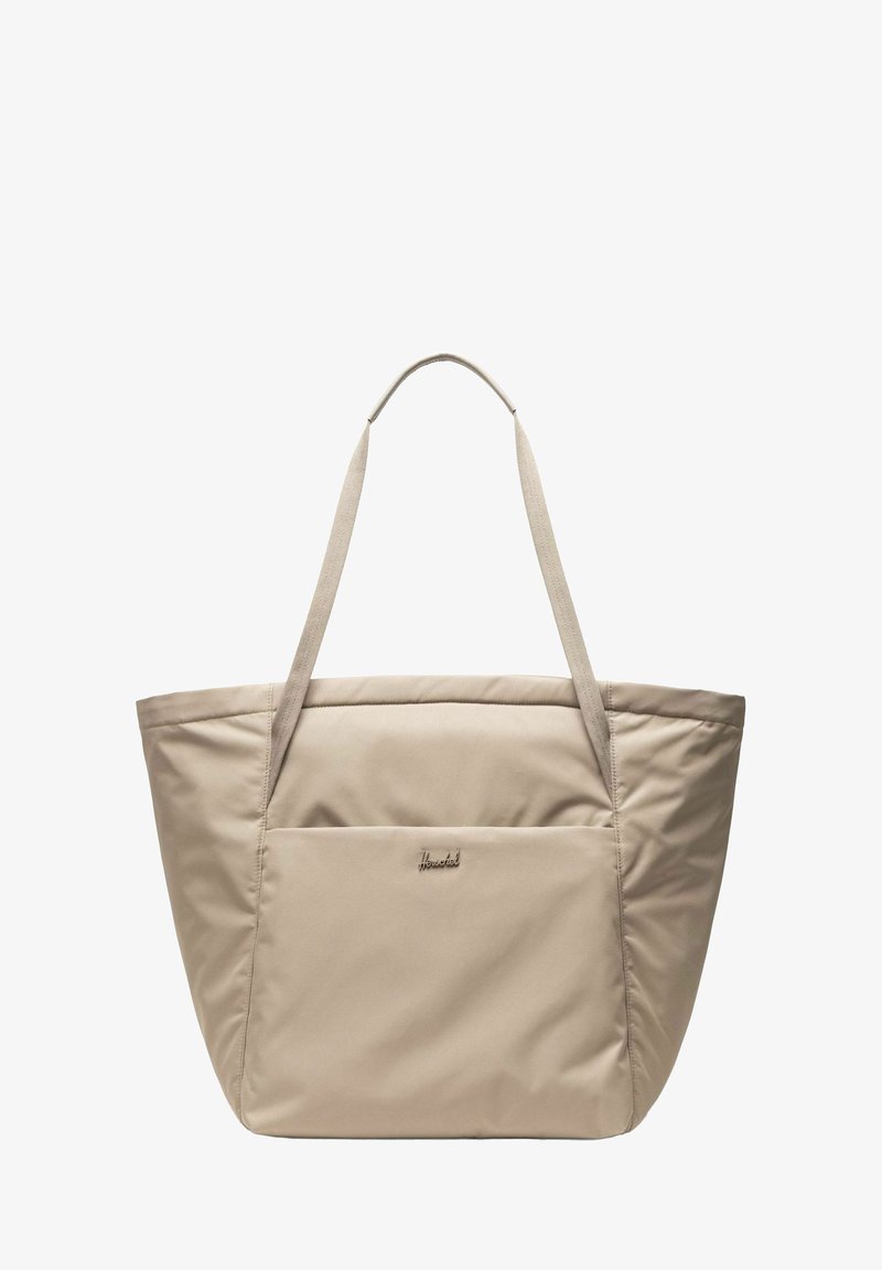 Sac fourre-tout en tissu doux beige avec de longues poignées et une poche extérieure avant, avec un petit détail de logo métallique.