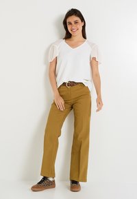 Top blanco de cuello en V con mangas de globo transparentes, combinado con pantalones anchos mostaza y un cinturón de estampado animal. Zapatillas marrones con acentos en negro y marrón claro.