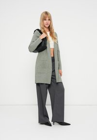 Απαλό πράσινο πλεκτό cardigan με δύο μπροστινές τσέπες, φορεμένο πάνω από μια λευκή μπλούζα crop top και συνδυασμένο με γκρίζες παντελόνες σε χαλαρή γραμμή και μαύρα μυτερά παπούτσια.