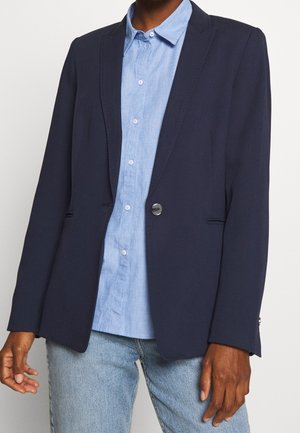 Personne portant un blazer bleu marine sur une chemise boutonnée bleu clair et un jean bleu clair, debout devant un fond uni.