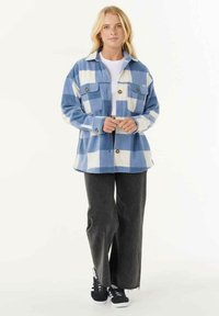 Blauw-witte gecontroleerde flanellen jas met twee borstzakken en metalen knopen, gecombineerd met zwarte wijde broek en zwarte sneakers.