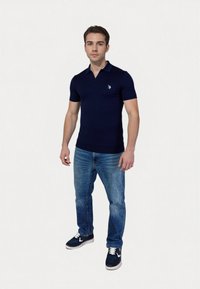 Polo de manga corta azul marino con cuello, que presenta un pequeño emblema azul en el pecho. Se lleva con jeans azul claro y zapatillas oscuras.