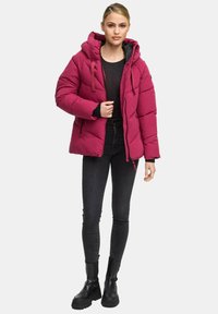 Vrouw in een helder roze pufferjack, zwarte top, donkere skinny jeans en zwarte chunky laarzen, staand tegen een effen achtergrond.