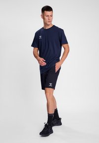 Marine bomulls t-skjorte med logo, kombinert med svarte shorts med logo. Modell i trening-sko, stående på en nøytral bakgrunn.