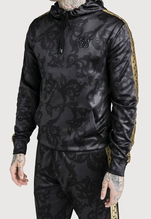 Schwarzer Hoodie und Hose mit dunklem Blumenmuster, goldenen Streifen an Ärmeln und Beinen sowie schwarzem, gesticktem Logo auf Brust und Oberschenkel.