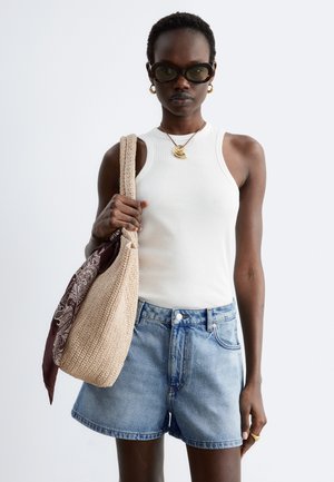 Femme portant des lunettes de soleil noires, un top blanc sans manches, un short en jean, un collier et des boucles d'oreilles en or, portant un sac tressé beige avec une écharpe marron.