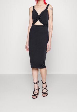 Vrouw met rood haar die een zwarte mouwloze midi-jurk draagt met een uitsnijding aan de voorkant en een gedraaide detail, gecombineerd met zwarte hoge hak sandalen met bandjes.