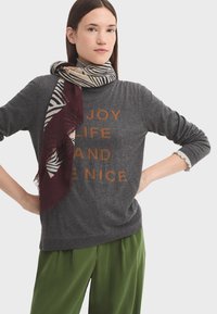 Grauer Pullover mit dem Text "FREUE DICH AM LEBEN UND SEI NETT" in Orange, kombiniert mit einem gemusterten Creme- und Marineblau-Schal und locker sitzenden grünen Hosen.