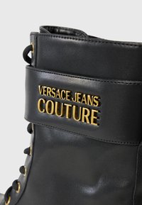 Stivale in pelle nera con logo dorato "Versace Jeans Couture", design con lacci e gambale imbottito, dotato di occhielli e dettagli di cucitura dorati.