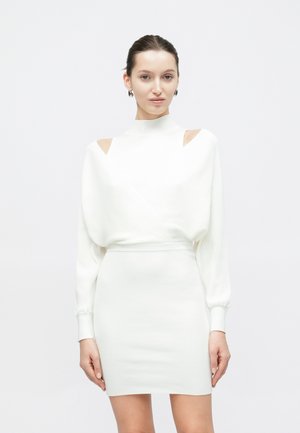 Donna che indossa una gonna bianca aderente e una maglia bianca a maniche lunghe con spalle scoperte, in piedi contro uno sfondo chiaro e uniforme.