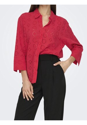 Femme portant une blouse rouge ample à œillets avec boutons et manches trois-quarts, avec un pantalon noir taille haute, une main dans la poche.