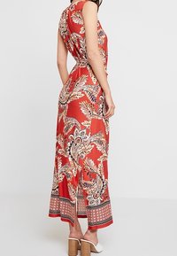 Abito lungo rosso in tessuto leggero con motivo paisley, schiena scoperta e spacchi laterali. Indossato con sandali con tacco bianchi.