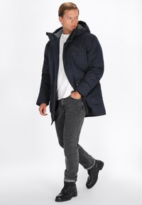 Marineblå pufferjakke med grå foret hætte, frontlynlås og sidelommer, båret over en hvid skjorte og mørke jeans, parret med sorte støvler.