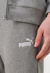 Pantalons de sport gris en tissu texturé, dotés d'une poche zippée et d'un logo Puma brodé blanc sur la cuisse. Gros plan sur le détail de la main.