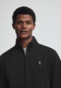 Giacca nera con zip e colletto alto, con un piccolo logo bianco. Tessuto morbido, design minimale e silhouette aderente.